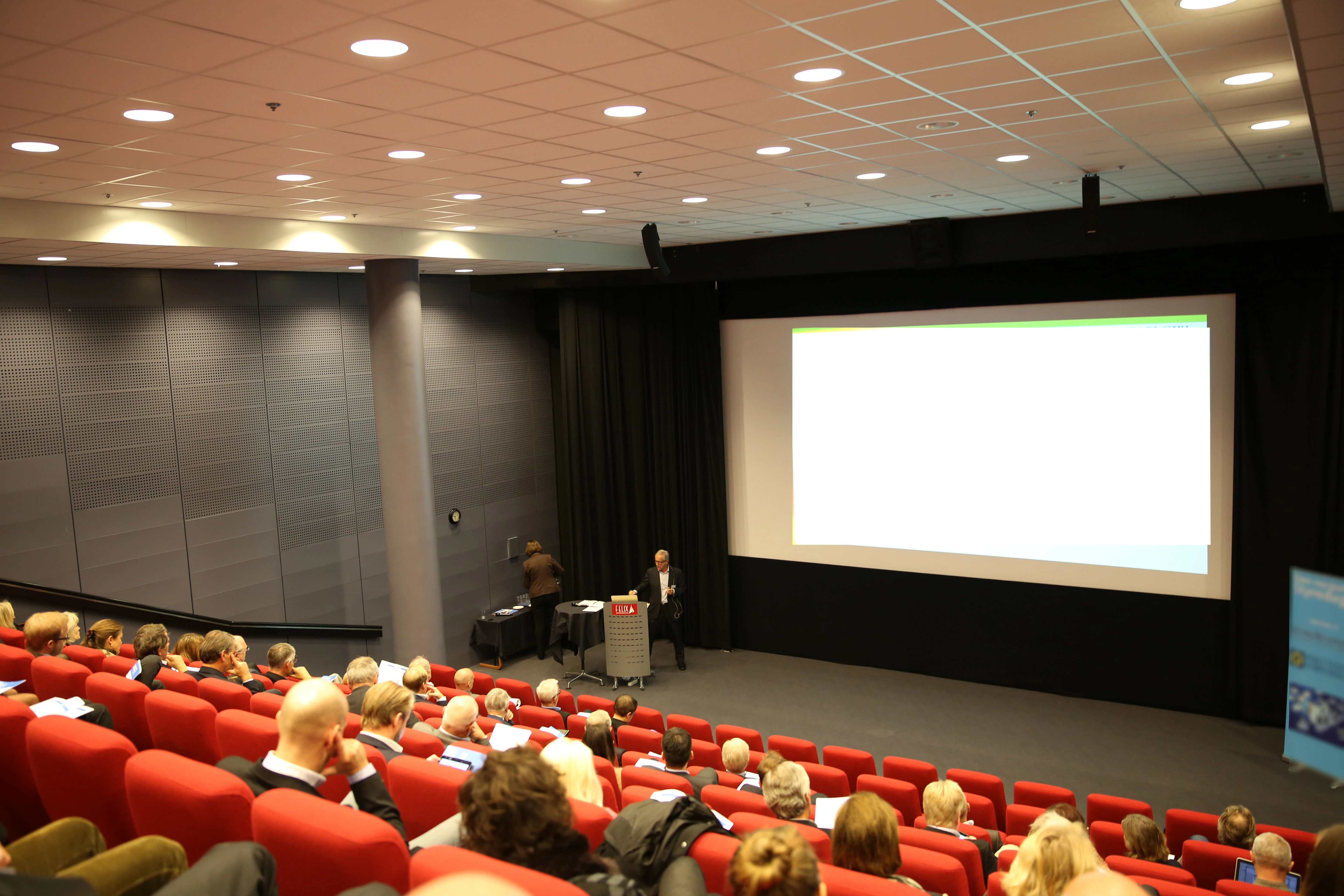 Conference speaker presenting to an audience in a theater with red seats and a large projection screen.