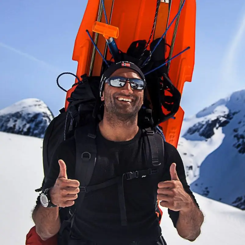 Man smiling and giving thumbs up while carrying skis on his back, standing in a snowy mountainous landscape under a clear blue sky.