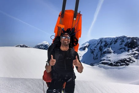 Smiling skier with gear on his back giving thumbs up in a snowy mountain landscape.