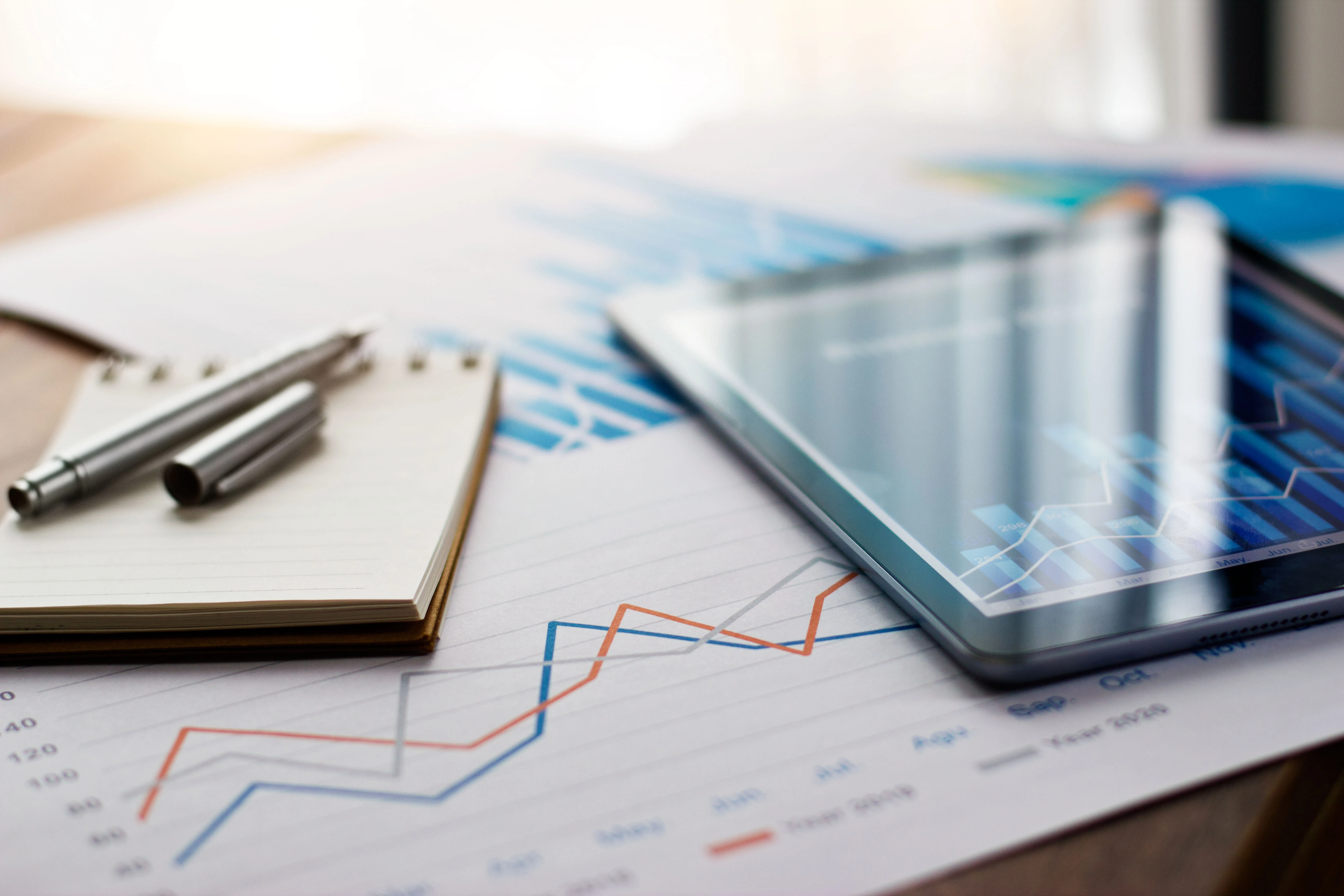 Tablet, notepad, and pen placed on top of financial charts with line graphs.