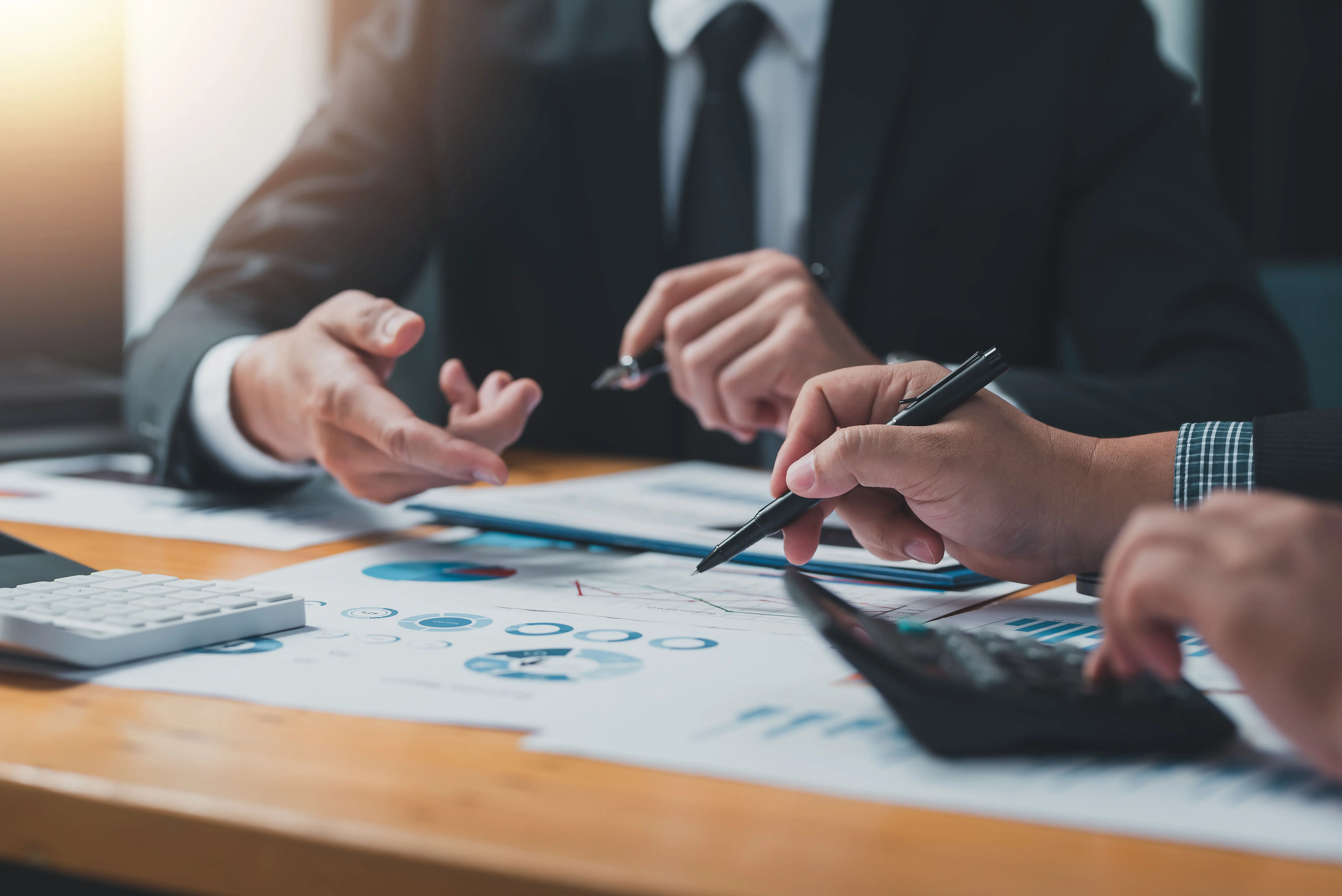 A group of business professionals in formal attire engaged in a meeting, reviewing financial charts and data on paper. One person is using a calculator, while others point to the documents with pens, indicating a discussion or decision-making process. The scene suggests a collaborative work environment focused on financial analysis or strategy planning.