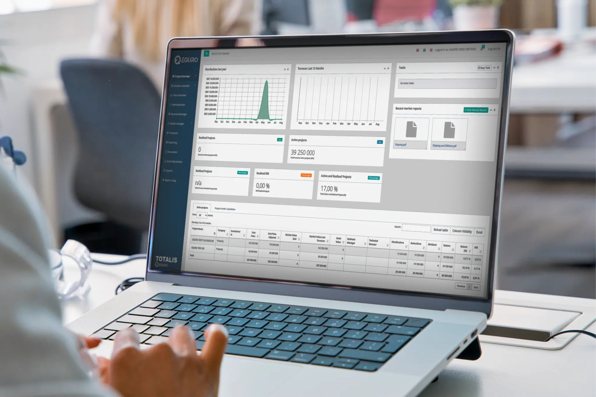 A person using a laptop with a dashboard displaying financial data, including charts for turnover, active projects, tasks, and a detailed table of investment metrics. The interface appears to be for managing projects or investments.