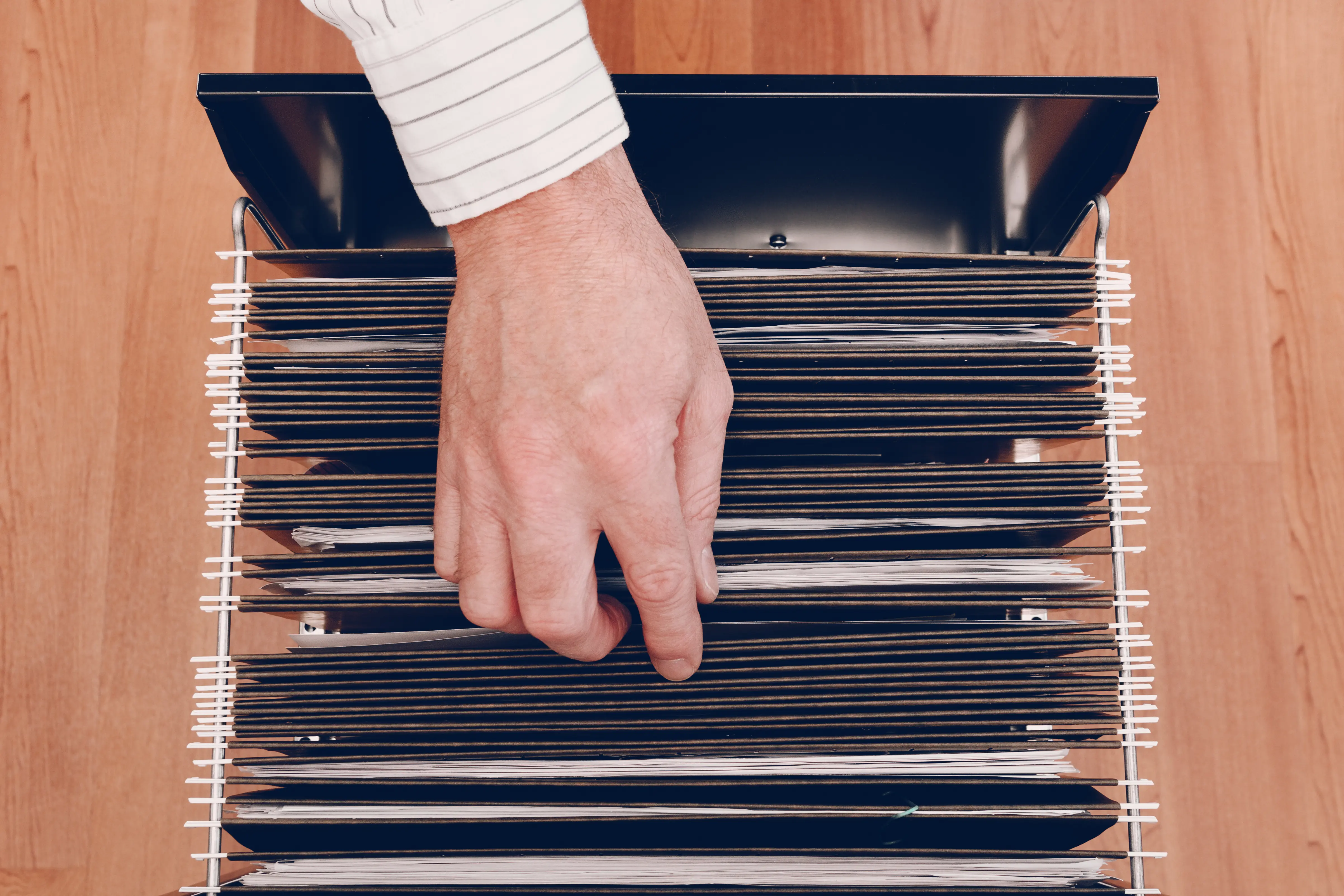 A person organizing or searching through a filing cabinet, with their hand pulling out a file from a row of folders. The cabinet is filled with documents neatly arranged in hanging file folders, suggesting a focus on paperwork, record-keeping, or data management in an office setting.