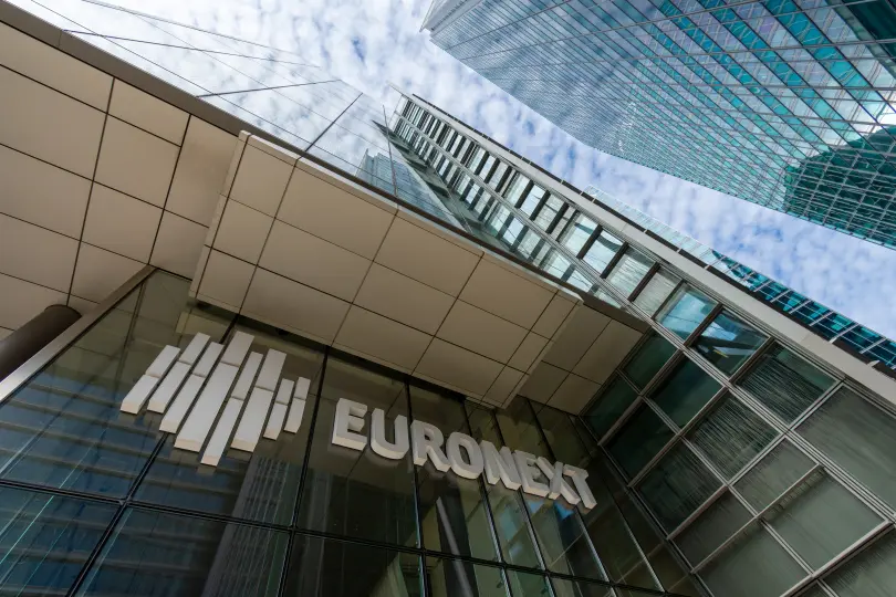 A low-angle shot of the Euronext building, with its logo prominently displayed on the glass facade. The modern skyscraper rises towards the sky, reflecting surrounding high-rise buildings, with a backdrop of a partly cloudy sky. The image conveys a sense of corporate scale and financial influence, representing Euronext as a major financial exchange.