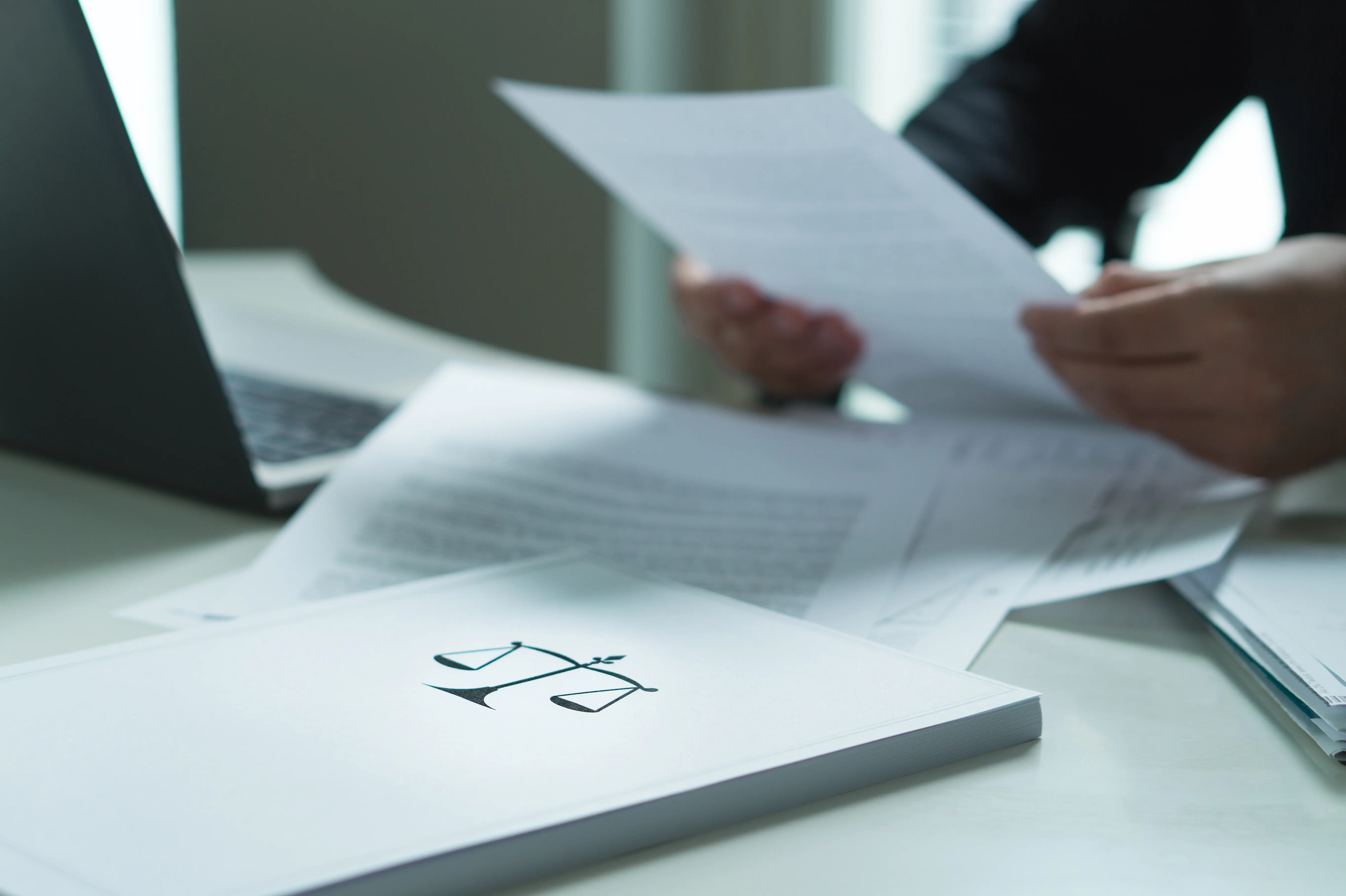 A person reviewing legal documents at a desk, with papers spread out and a folder featuring a scales of justice symbol. A laptop is partially visible in the background, suggesting a professional or legal setting.